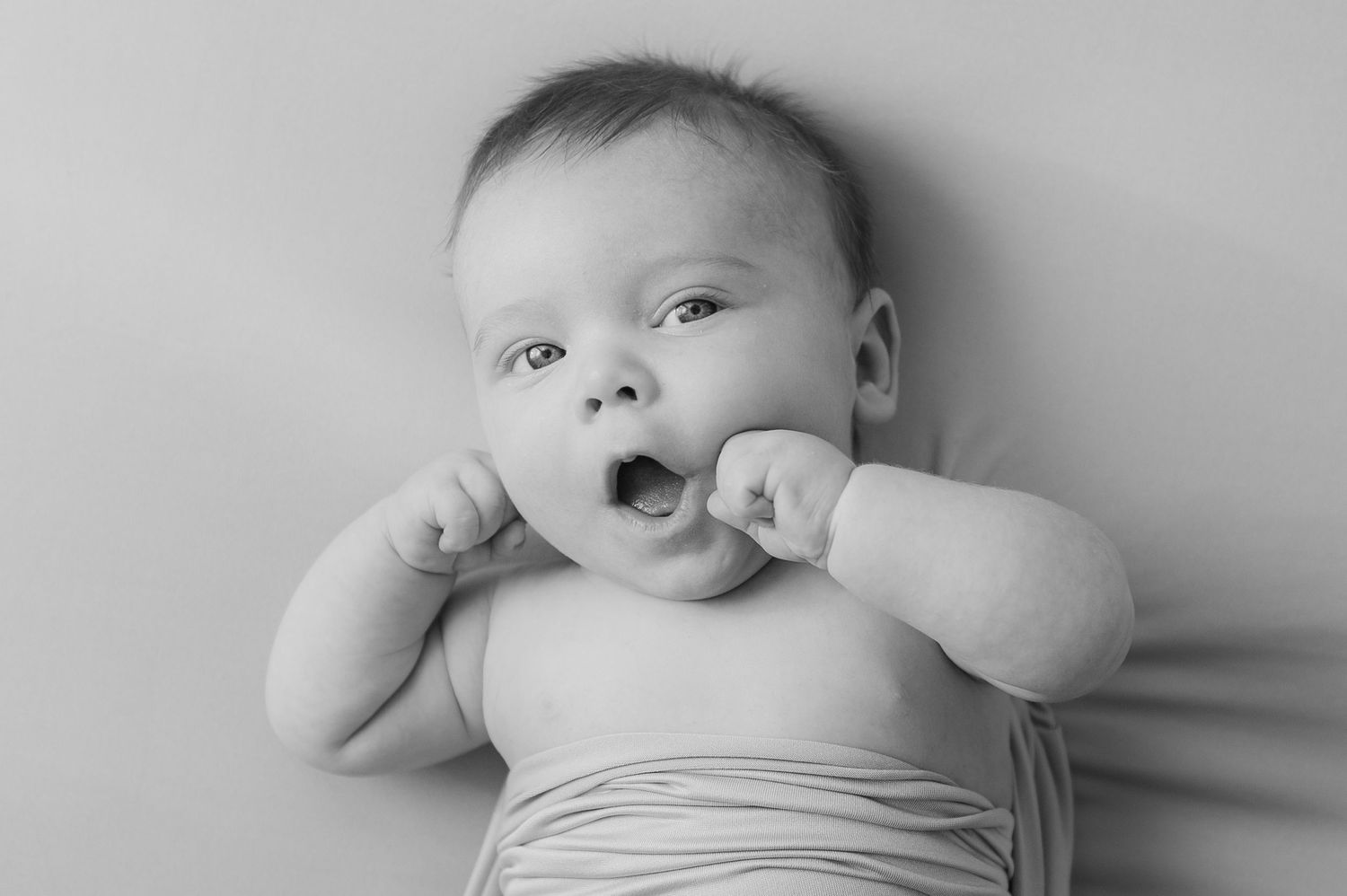 Black and white portrait of a yawning baby lying down with tiny fists raised near their face.