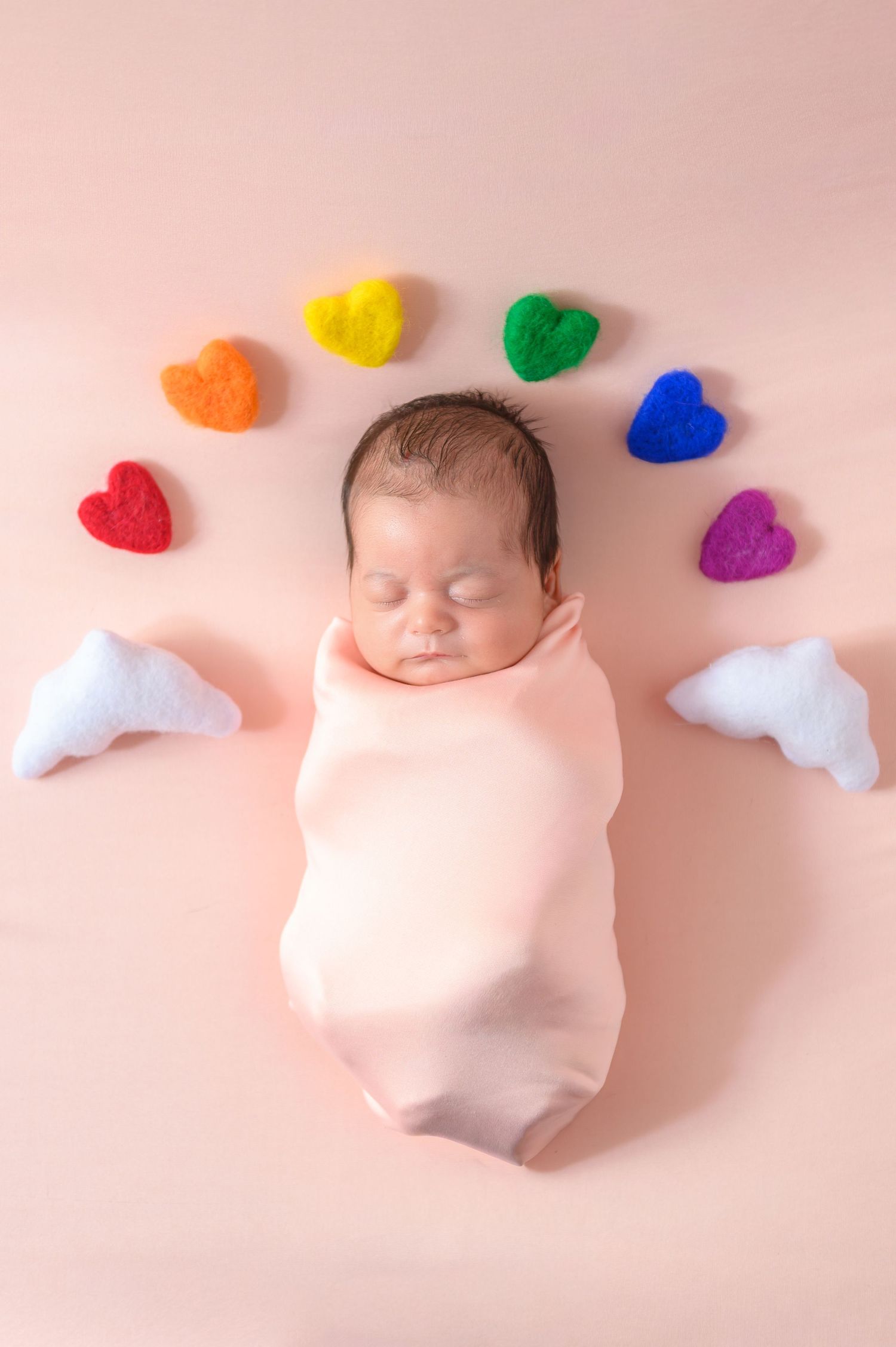 Sleeping newborn swaddled in pink with colorful heart-shaped decorations and white cloud shapes arranged in rainbow order.