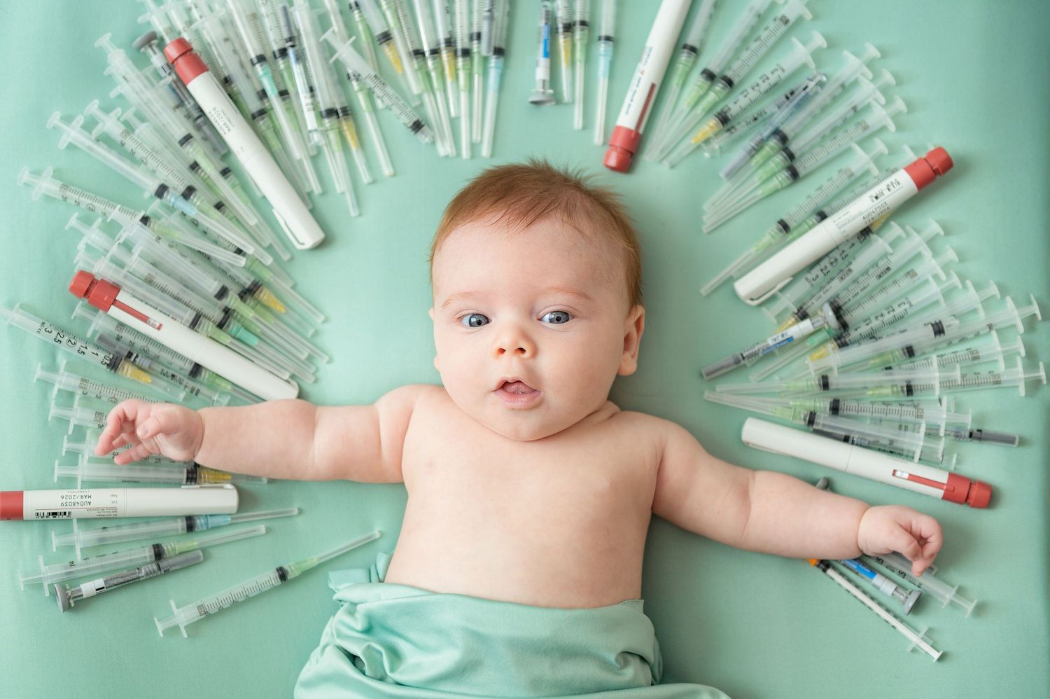 A baby lies surrounded by arranged IVF medical syringes on a mint green background in an artistic composition.