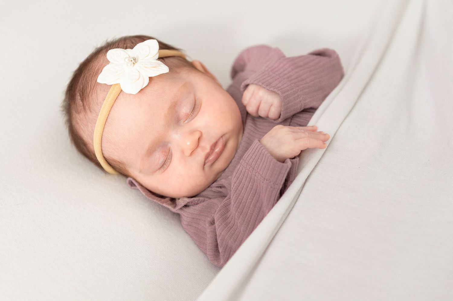 A sleeping newborn baby wearing a mauve outfit with a white bow headband rests peacefully in a collage of four photos.
