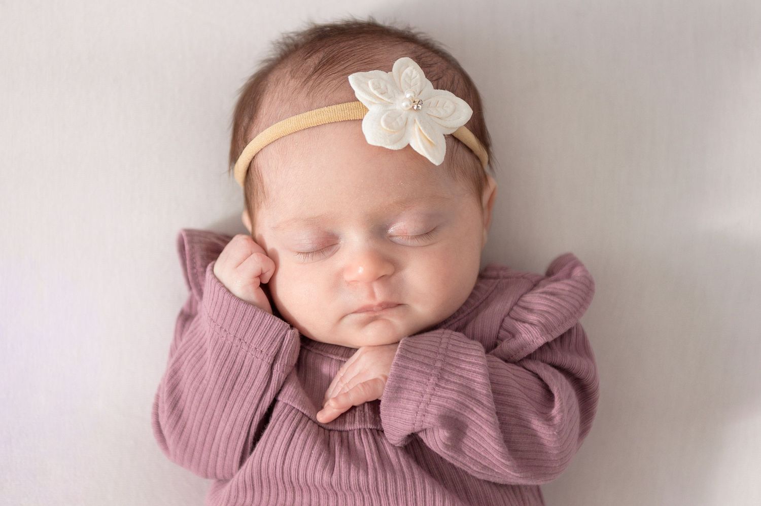 Sleeping baby wearing mauve knit sweater and white bow headband rests peacefully on soft blanket.