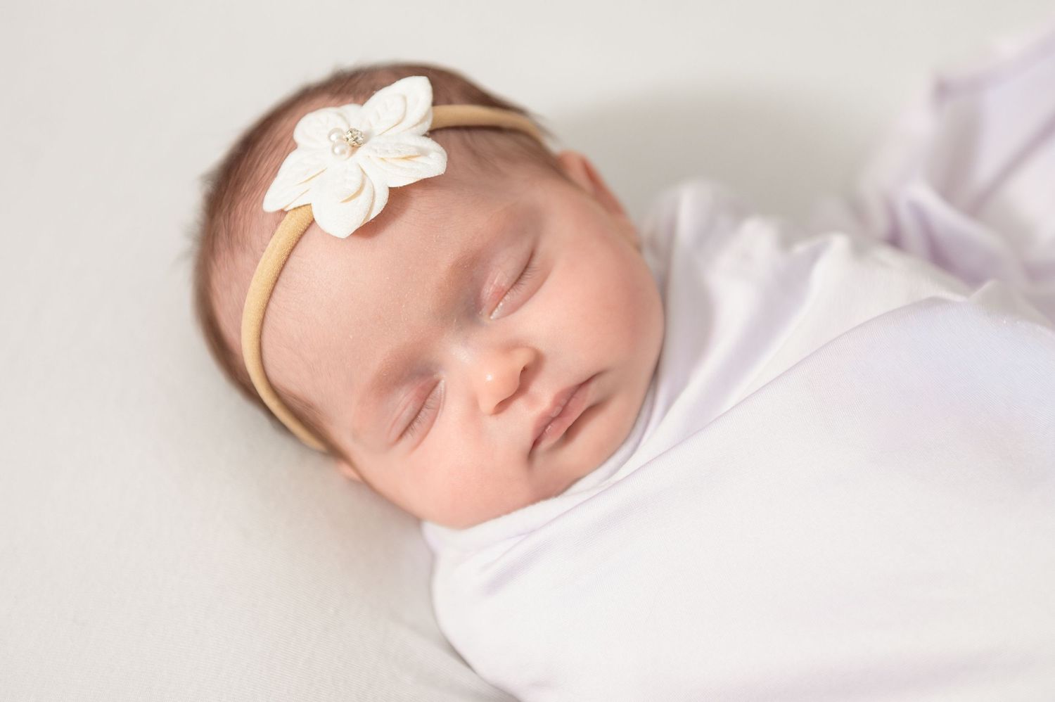 A sleeping baby wearing white lies peacefully on soft white bedding with a delicate bow hair accessory.