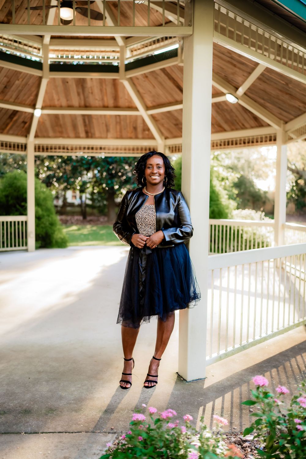 Woman in navy tulle skirt and leather jacket smiling under a gazebo surrounded by flowers and greenery.