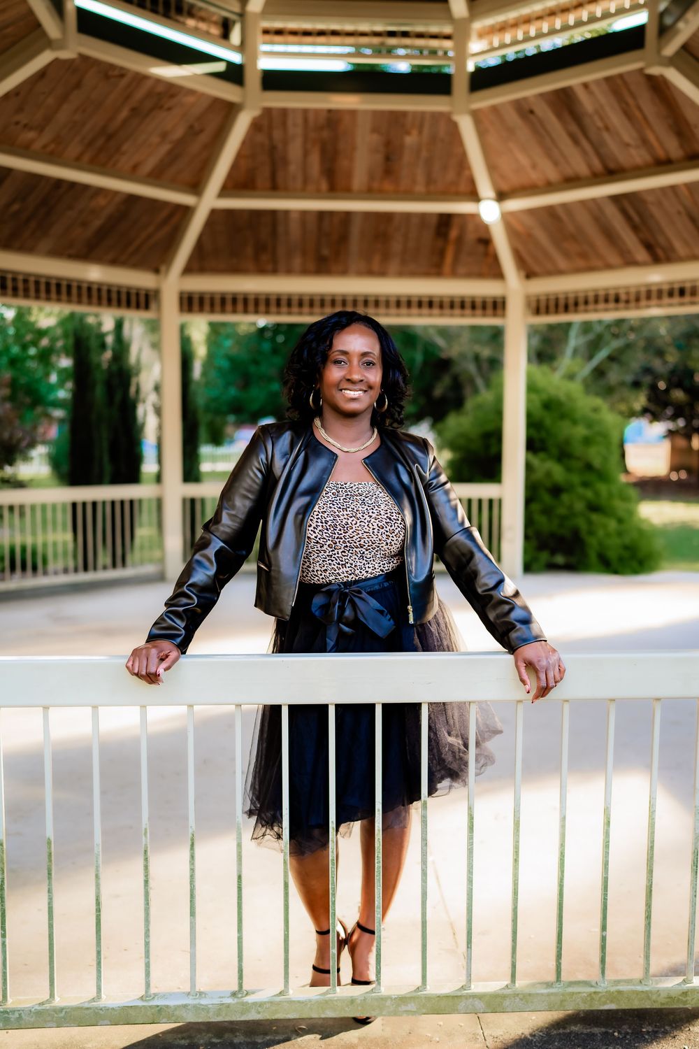 A smiling woman in a leather jacket and skirt poses at a wooden gazebo railing in a sunny park.