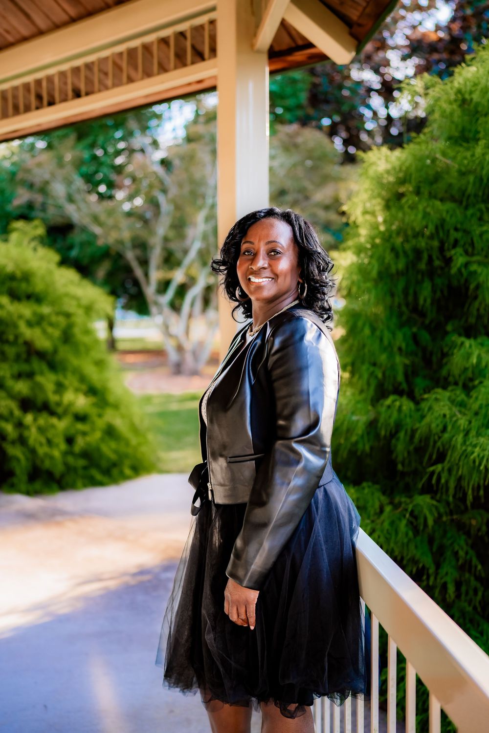 Smiling woman in black leather jacket and tulle skirt poses on a porch surrounded by lush green trees.