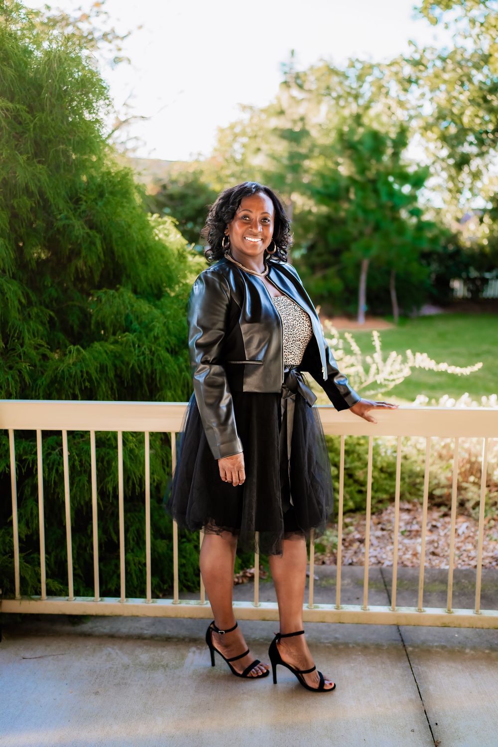 A smiling woman in a black tulle skirt, leather jacket, and heels poses on an outdoor patio surrounded by lush greenery.
