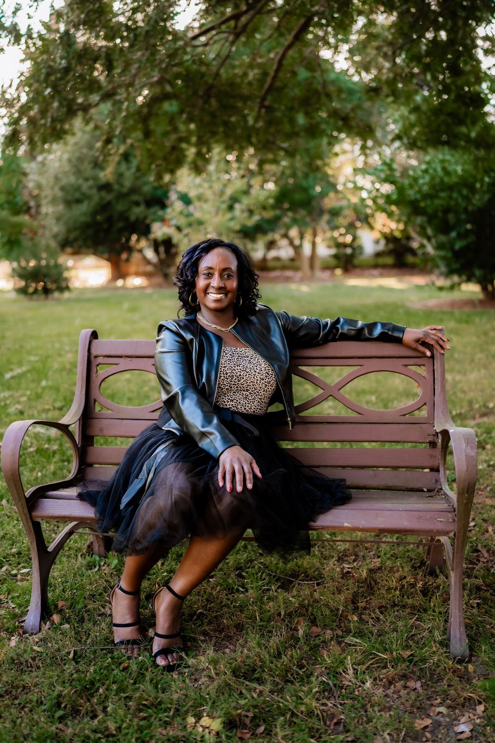 Smiling woman in leather jacket and tulle skirt sitting on a park bench surrounded by lush green trees.