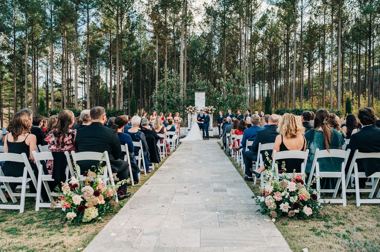Panoramic view of an outdoor wedding ceremony in a pine forest with white chairs and guests seated in rows.