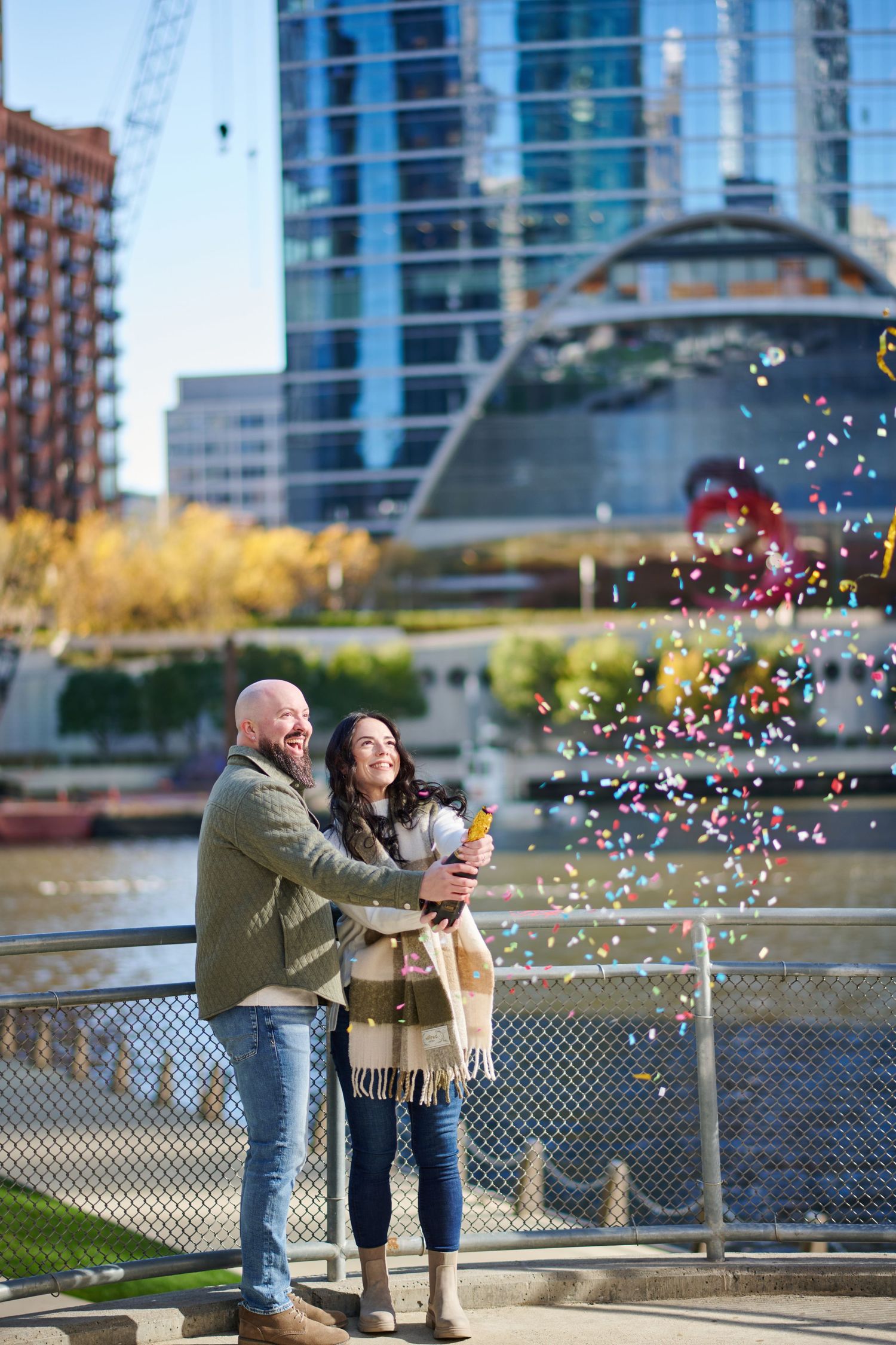 A couple celebrates with confetti by a waterfront in downtown Chicago with modern buildings and fall foliage in background.