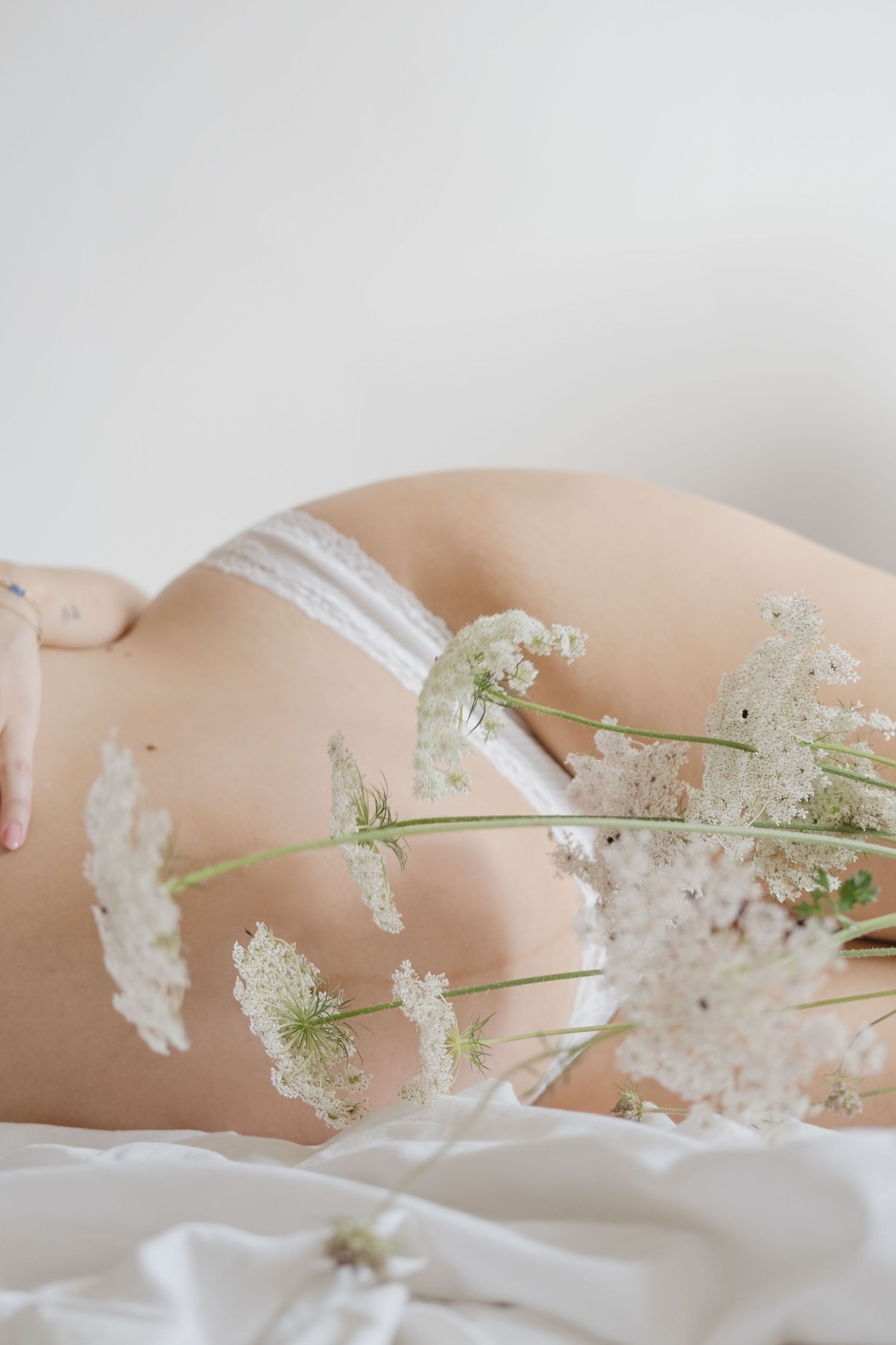 Delicadas flores blancas campestres dispuestas artísticamente sobre la barriga de una embarazada tumbada con una iluminación suave..