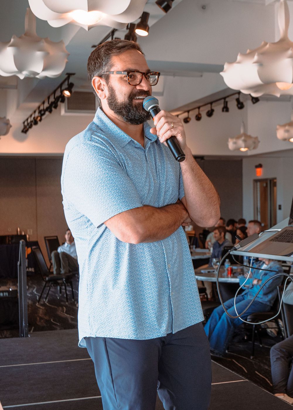 A speaker in a light blue polo shirt addresses an audience in a modern office or conference space.