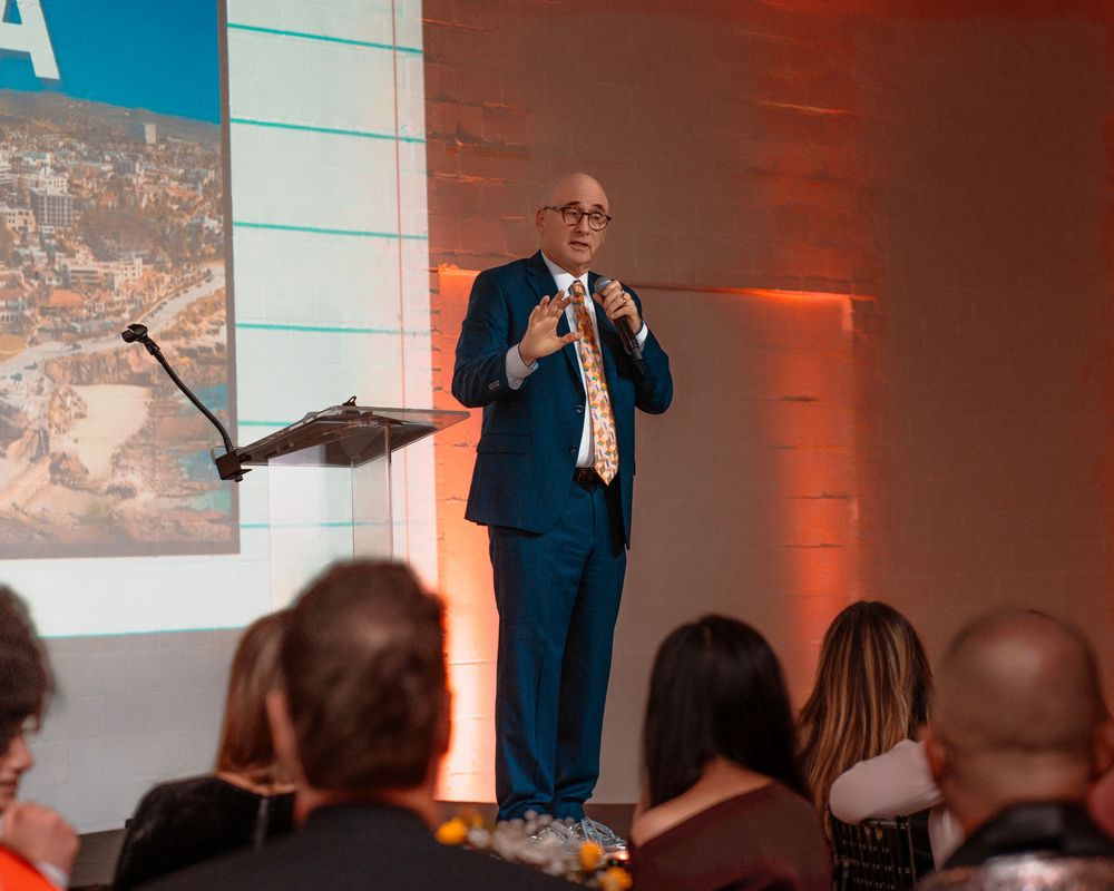 Speaker in a navy suit gestures while giving a presentation in front of a projected screen with orange lighting.