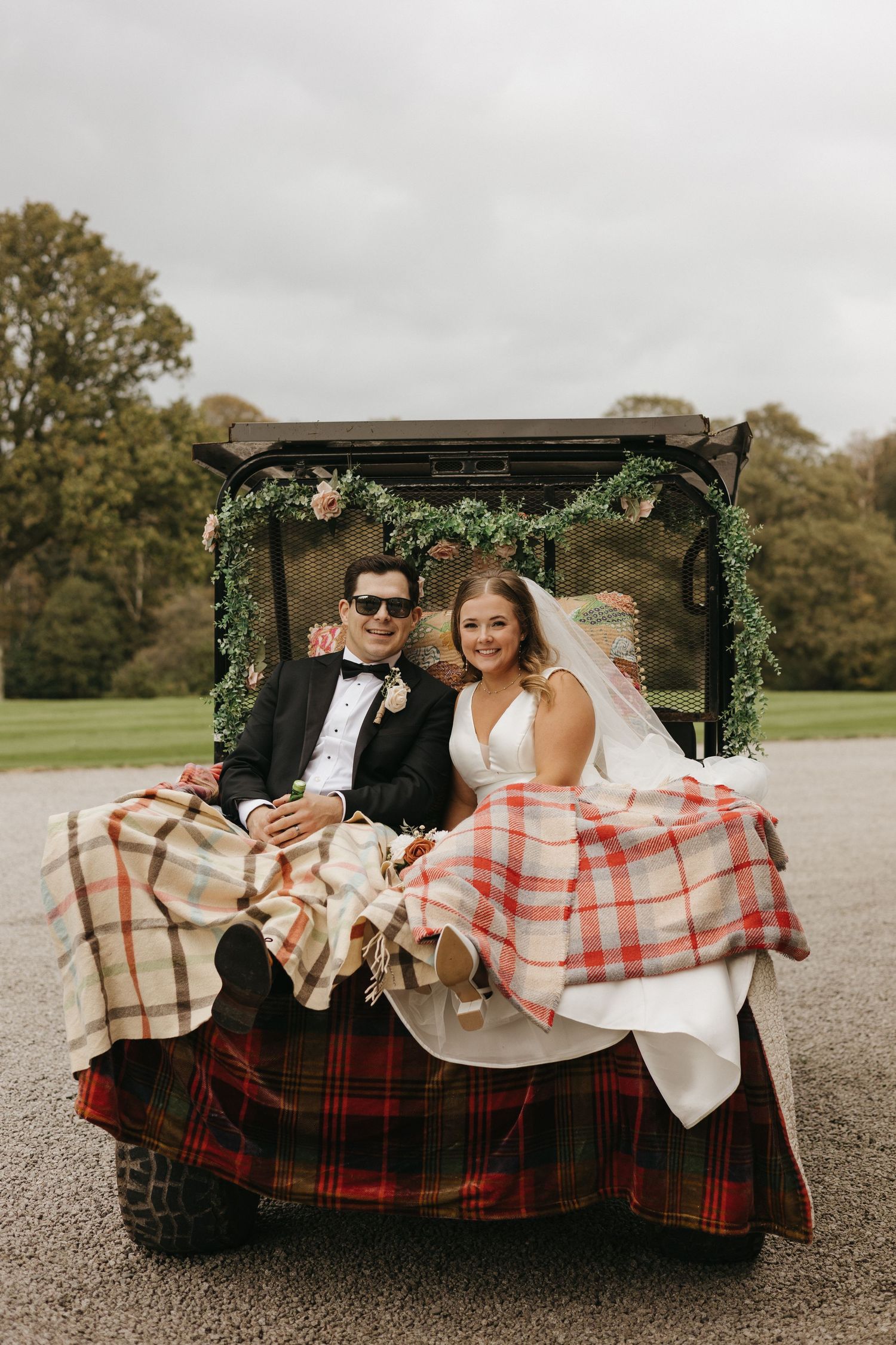 Wedding couple relaxes in vintage golf cart decorated with plaid blankets and greenery garland.