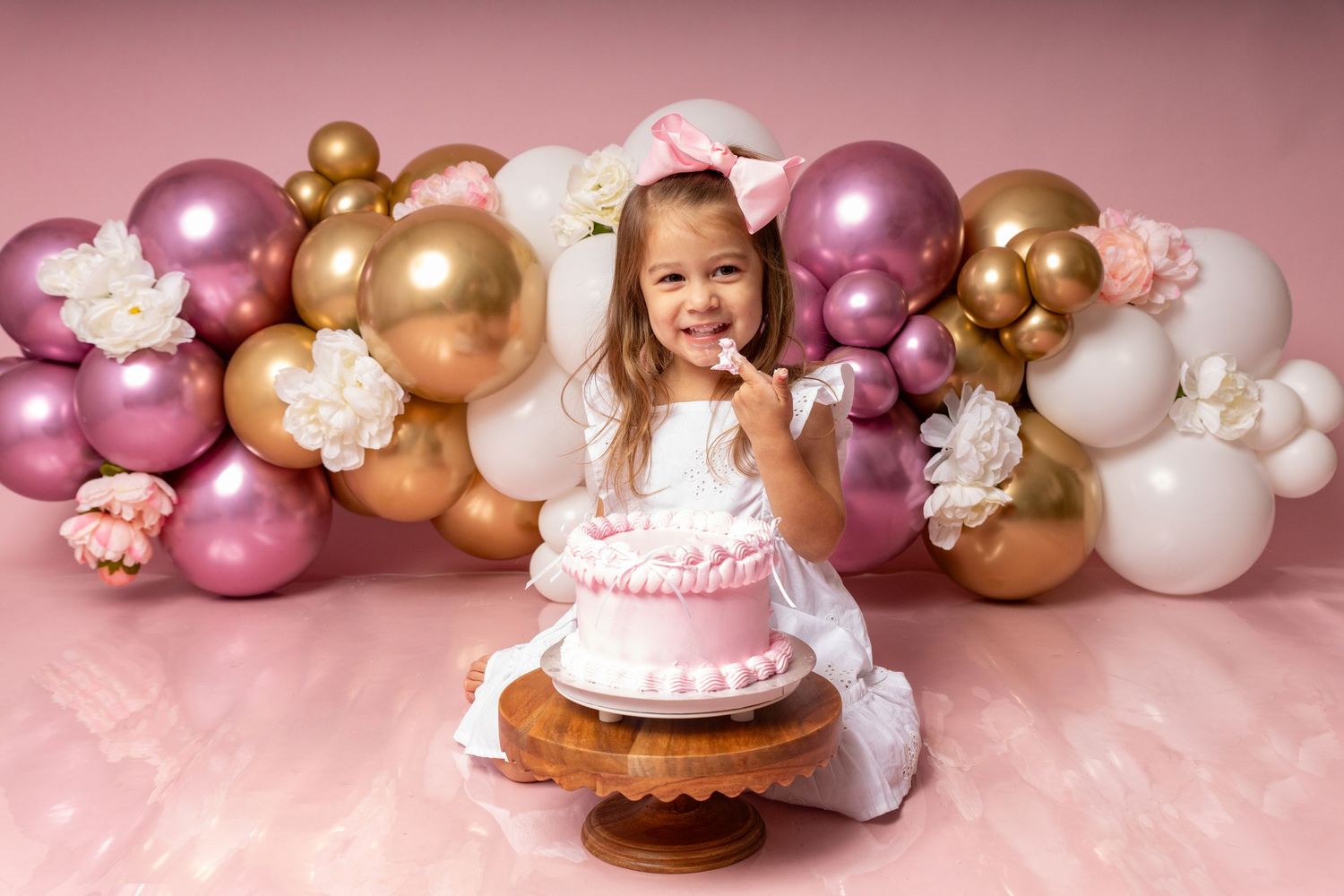 Baby wearing pink bow smiles with cake surrounded by colorful flower wall decoration.