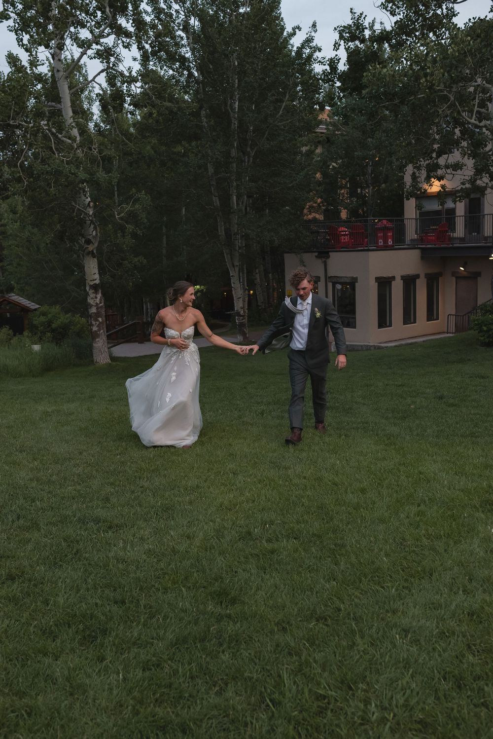 A couple in wedding attire walking and playing together on a lush green lawn surrounded by trees at dusk.