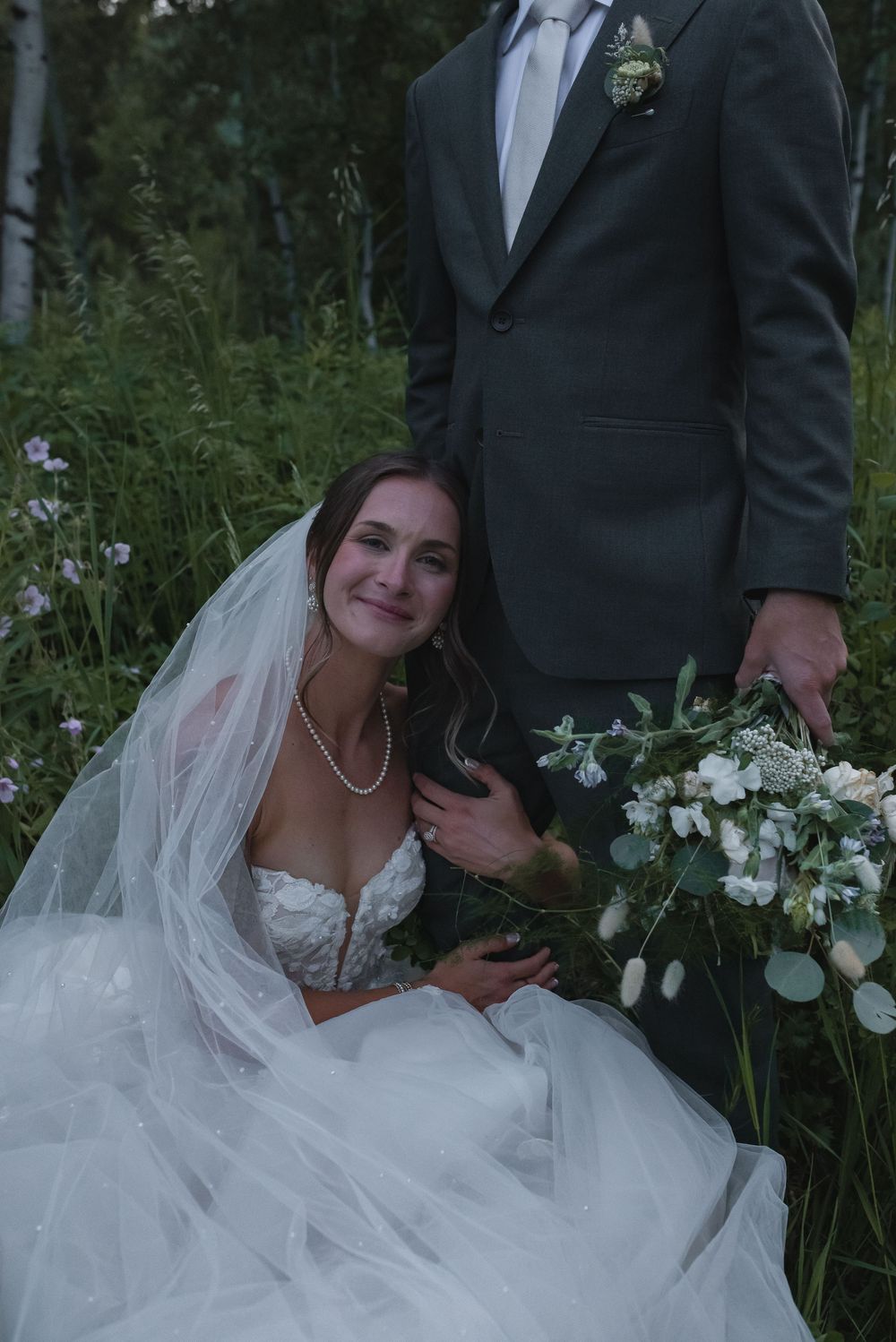A bride and groom pose together in a garden with white flowers and greenery in the background.