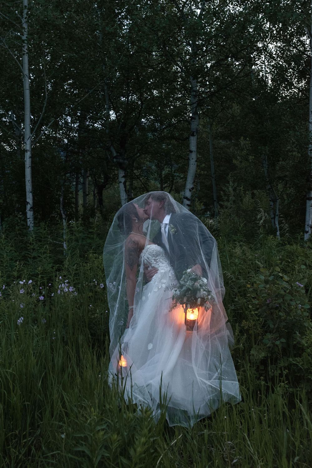 A romantic wedding couple sits in tall grass at dusk with glowing lanterns creating a dreamy atmosphere.