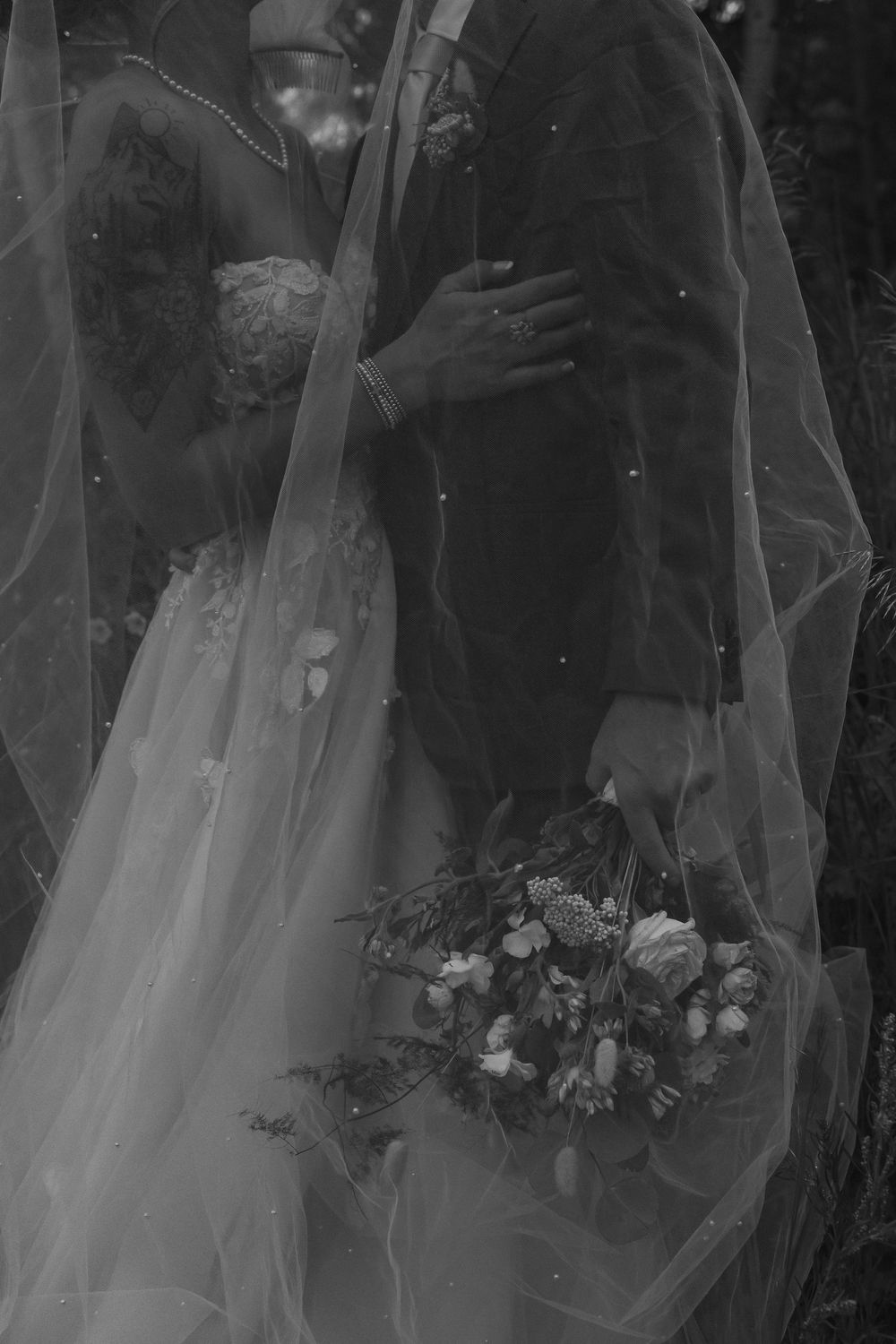 A black and white wedding photograph showing a couple embracing in formal attire with a flowing veil and bouquet.