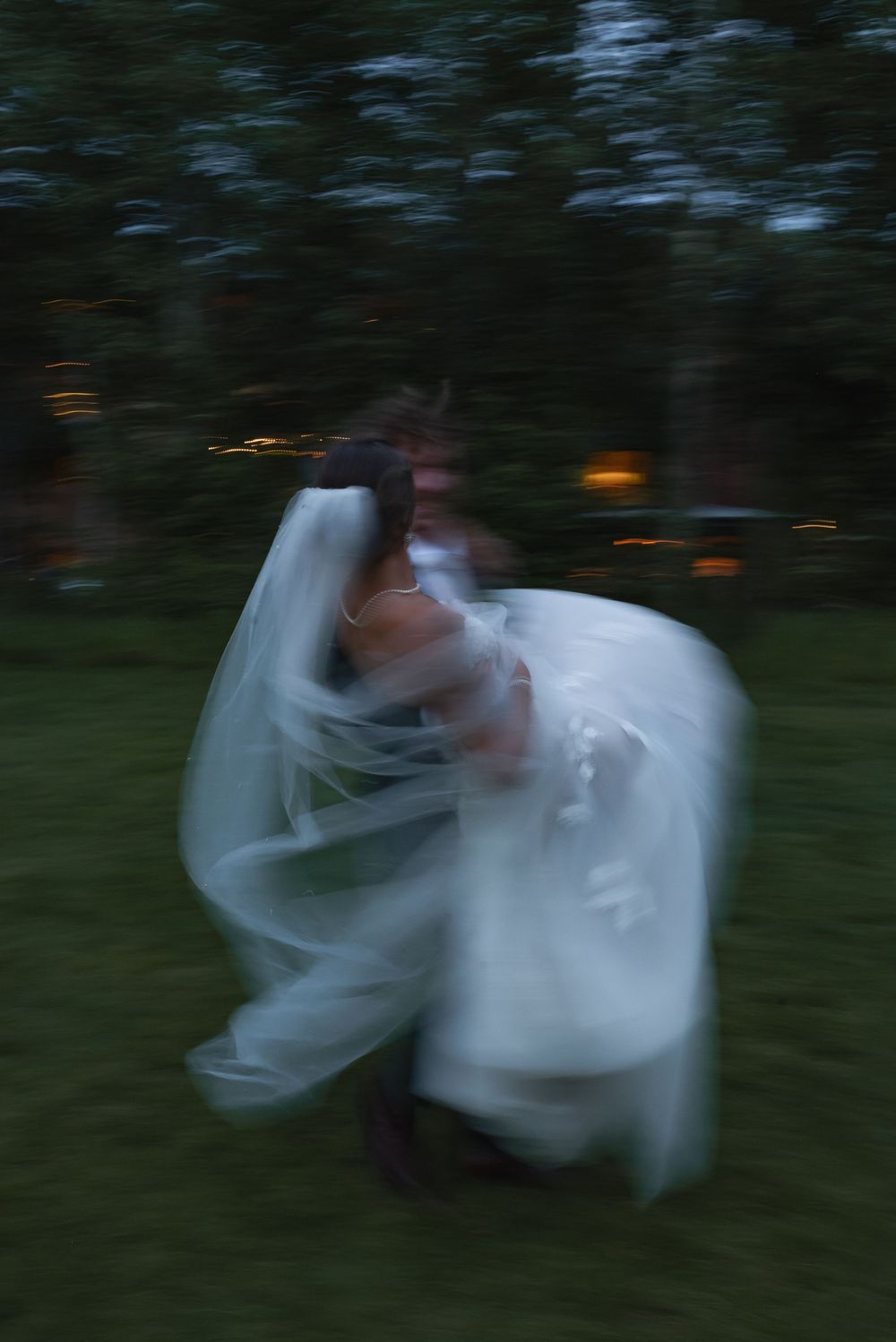 Motion-blurred figure in a flowing white wedding dress spins in a dark garden setting at dusk.