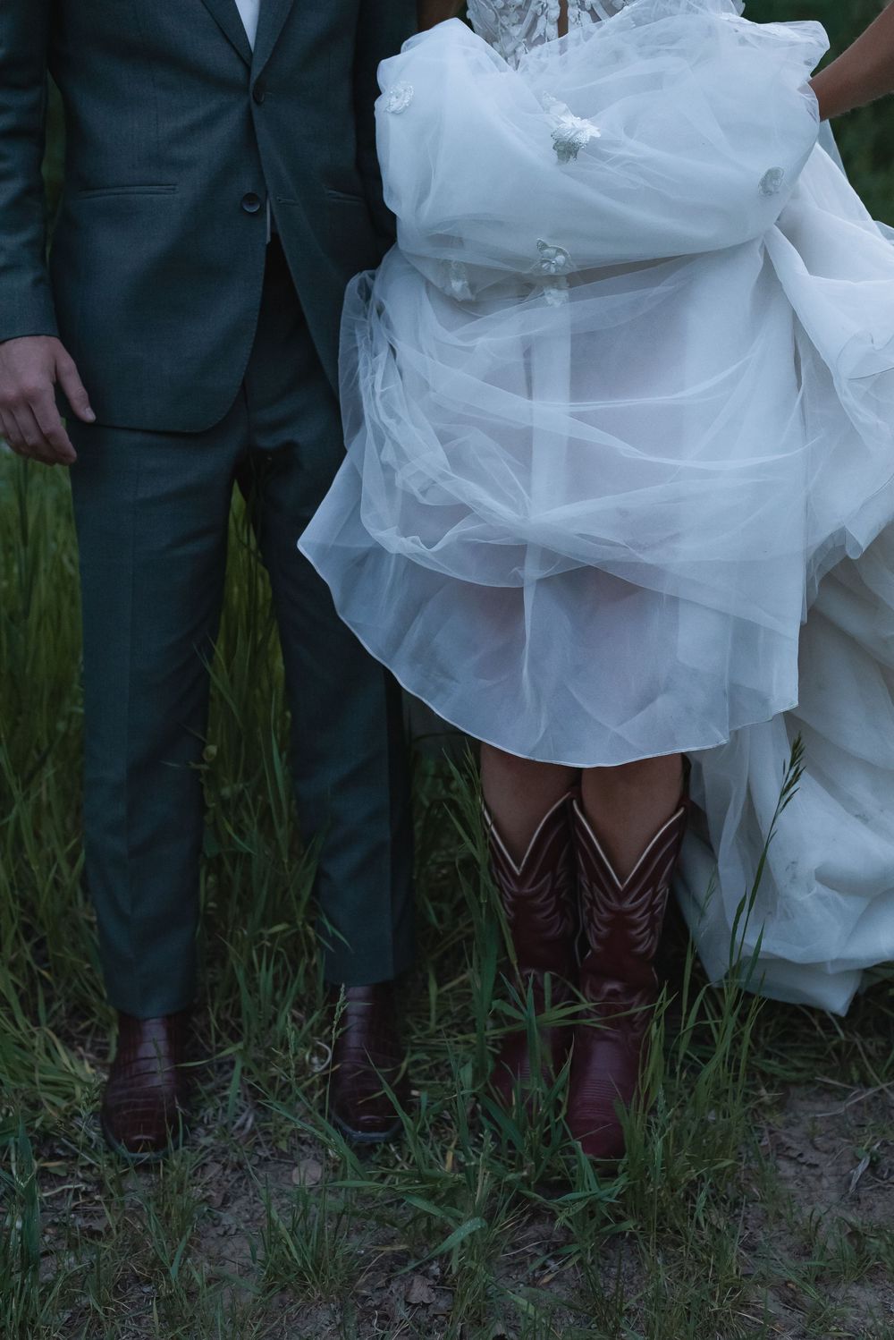 Wedding party members wearing cowboy boots and formal attire stand together in tall grass.