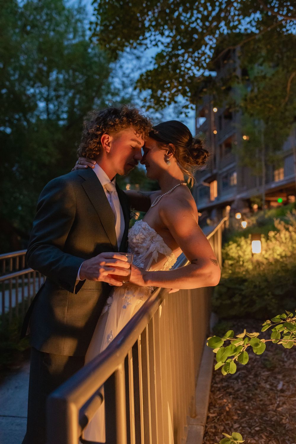 A couple shares an intimate moment on a porch railing at dusk with warm lighting and trees in the background.