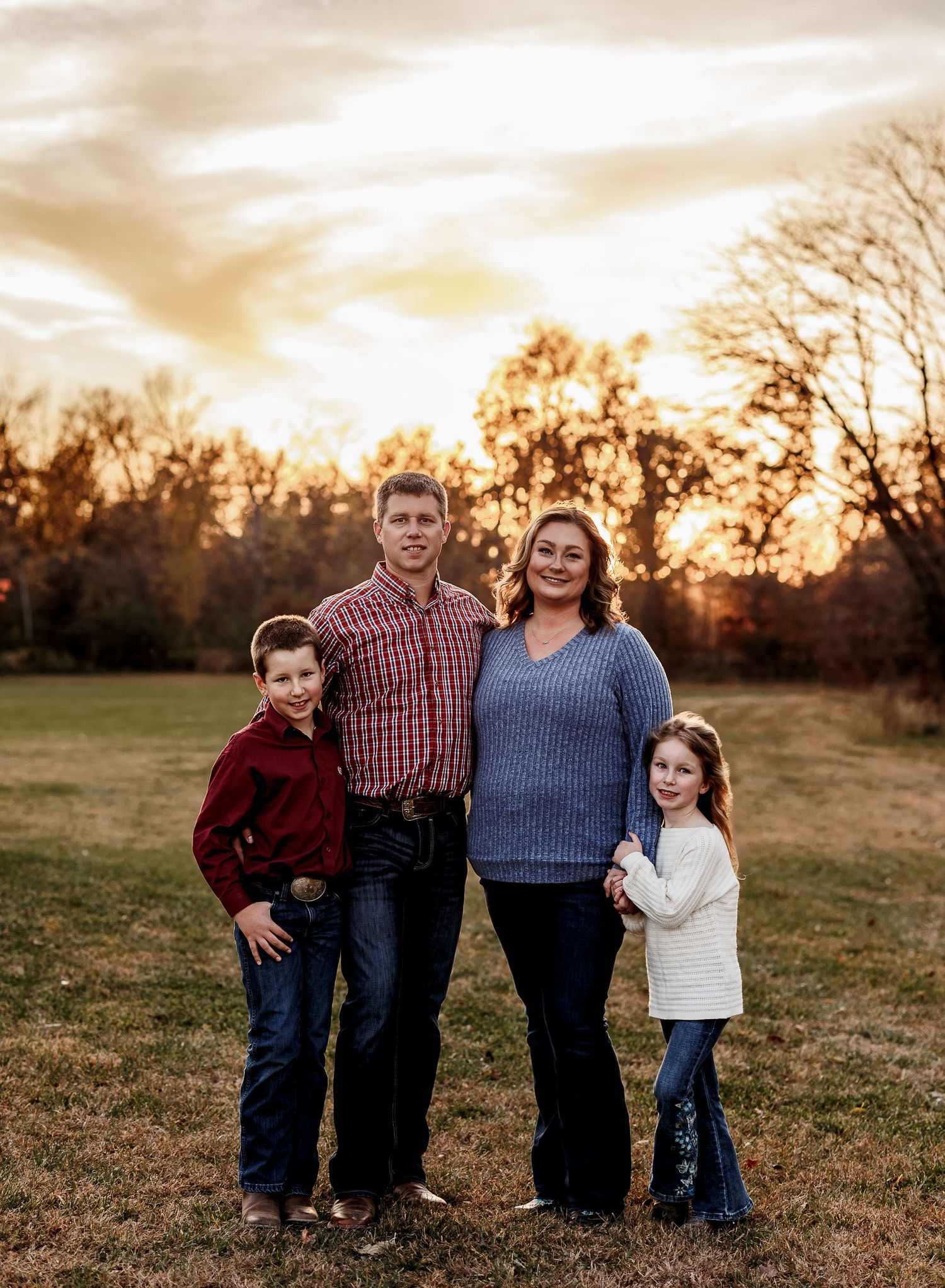 Family of four poses together in an open field at sunset with warm golden lighting and natural backdrop.