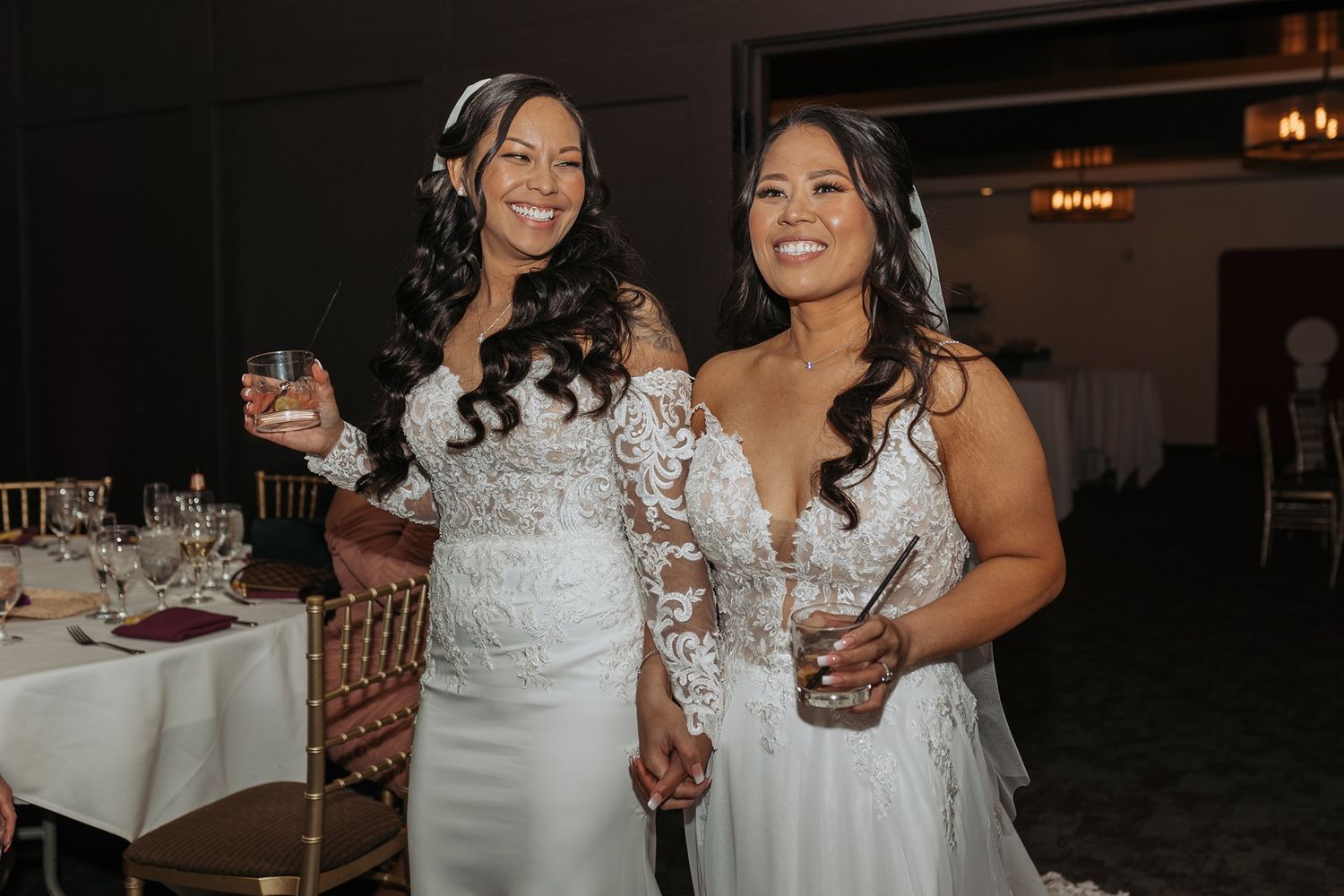 Two brides enter together holding 2 cocktails at a wedding reception in an elegant indoor venue.