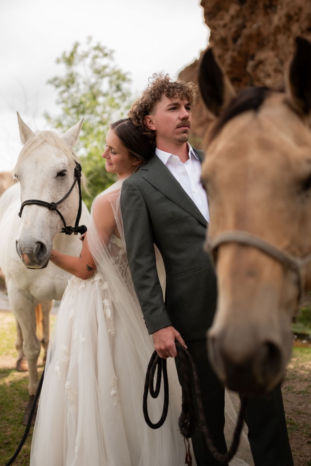 A couple in wedding attire poses with a white horse and donkey in a rustic outdoor setting.