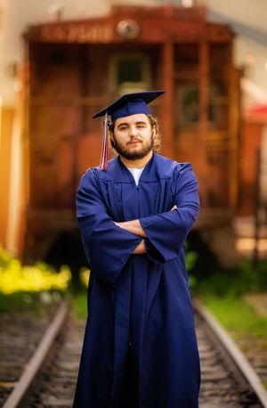 Graduate in blue cap and gown poses on railroad tracks for graduation photos at sunset.