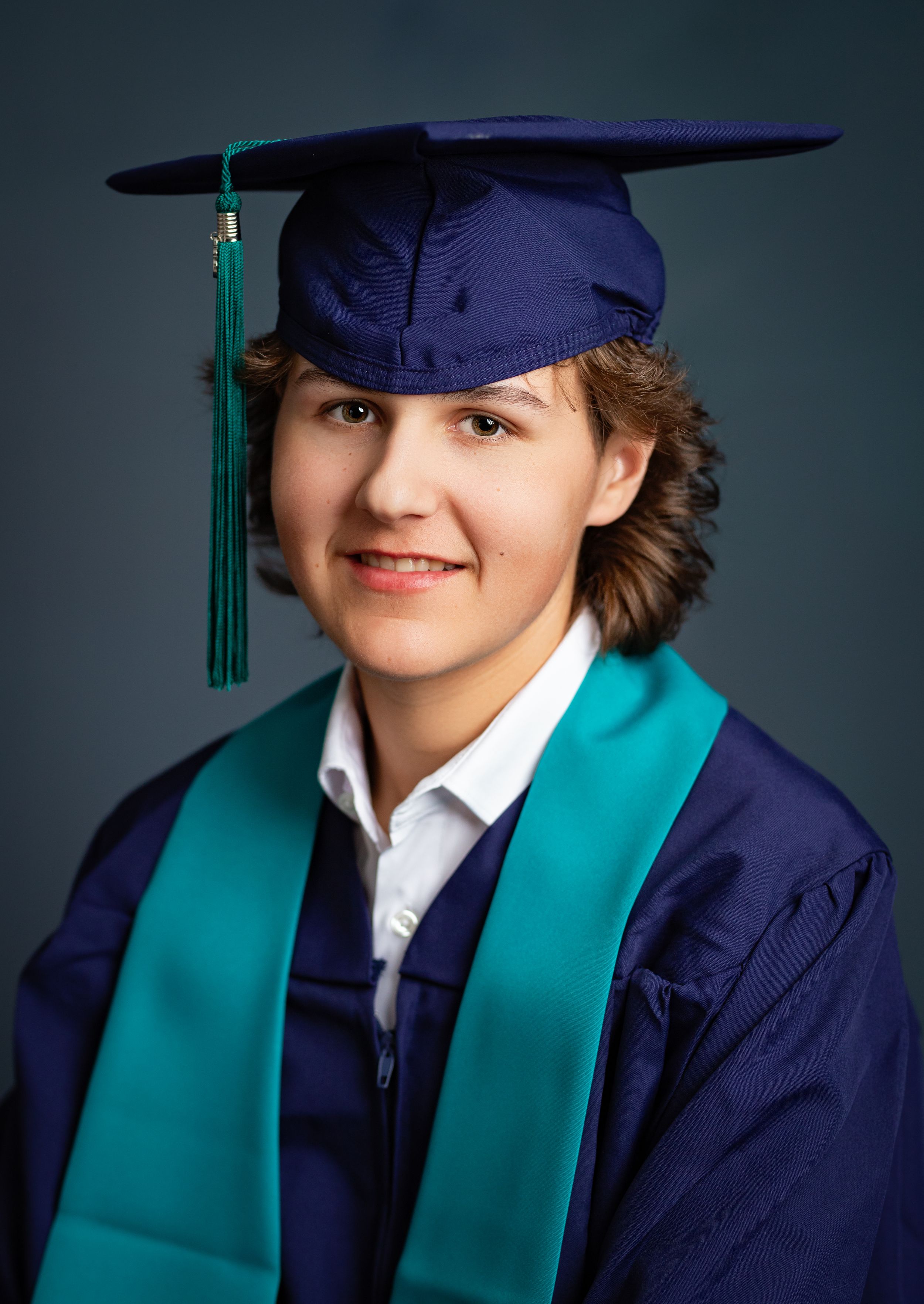 Graduate in navy blue cap and gown with turquoise stole poses for series of graduation portraits against dark background.