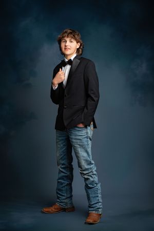 A person in a black blazer and blue jeans stands confidently against a dark moody backdrop.