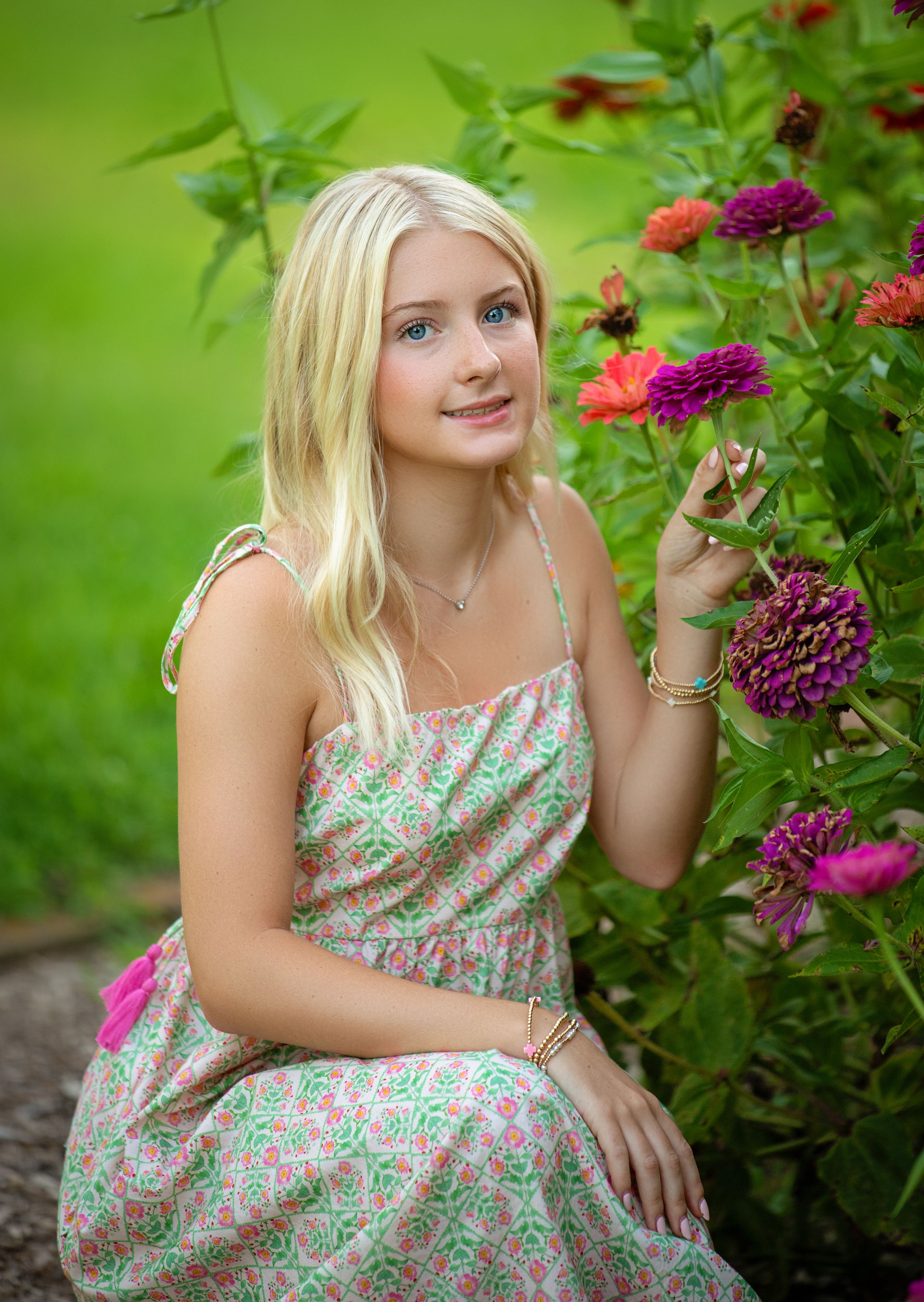 Blonde girl in floral sundress posing beside colorful zinnia flowers in a lush green garden.