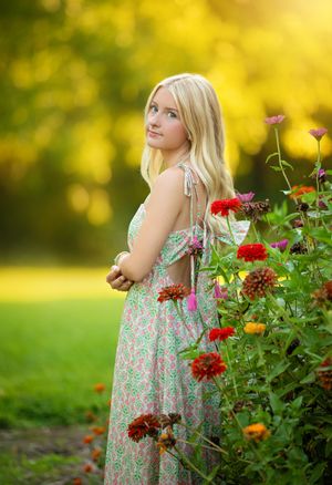 Blonde woman in floral sundress standing beside colorful zinnia flowers in a sunlit garden.