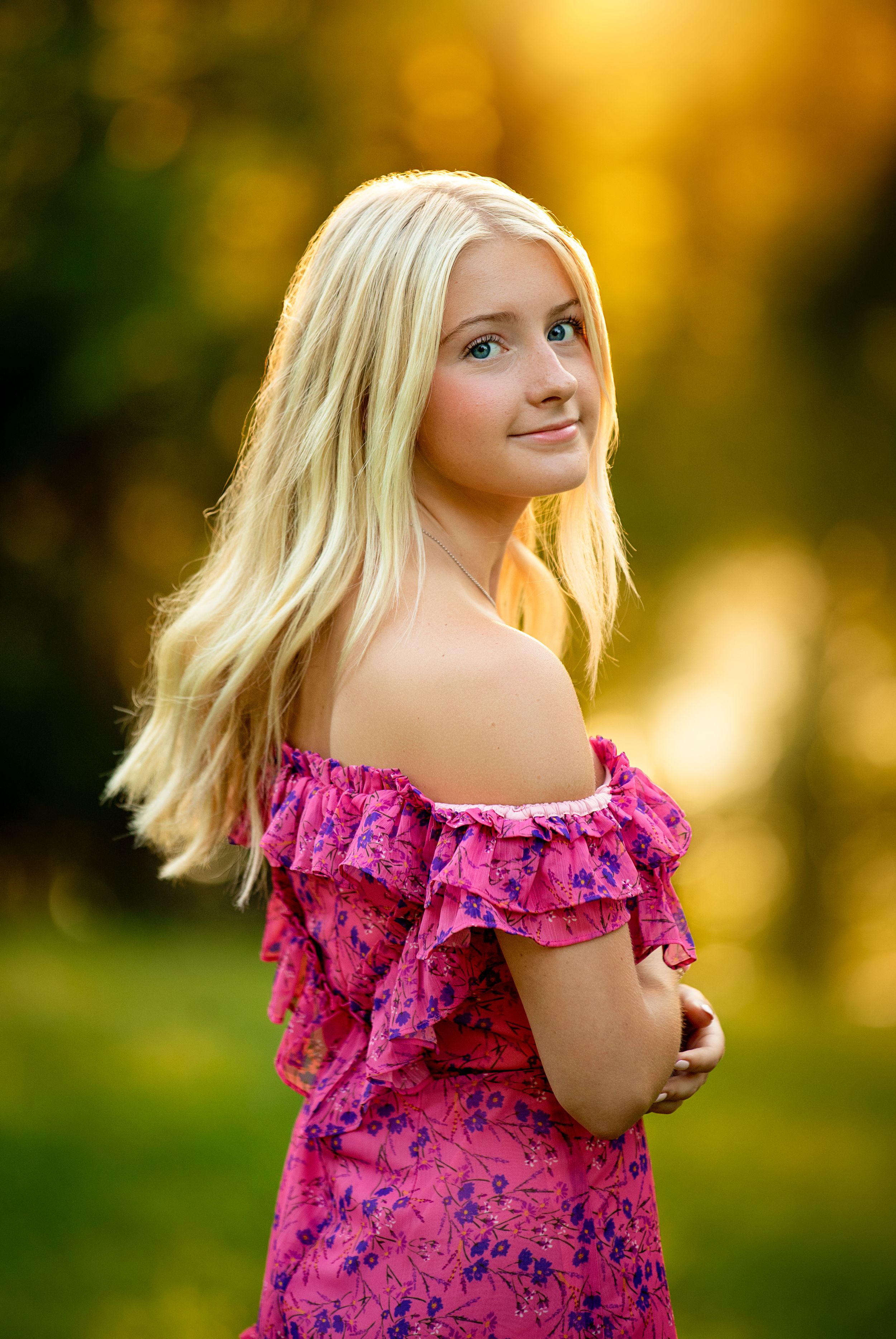 Young blonde girl in pink off-shoulder floral dress poses outdoors in golden sunset light.