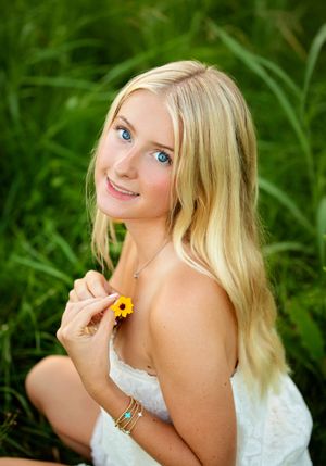 Smiling blonde girl with blue eyes holding a yellow flower, sitting in lush green grass wearing a white dress.