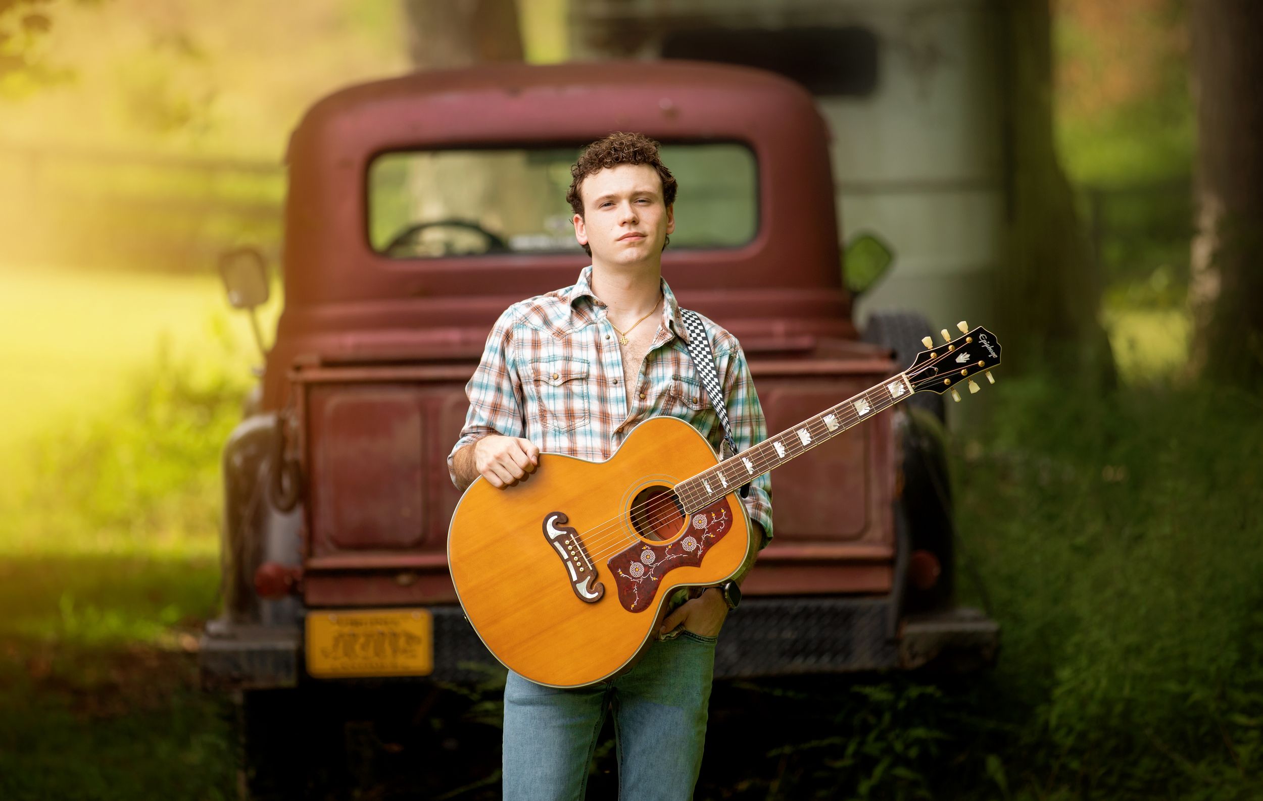 Country musician poses with acoustic guitar in front of vintage red pickup truck during golden hour photoshoot.