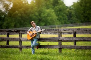 Musician in denim plays acoustic guitar while sitting on wooden fence in grassy field on sunny day.
