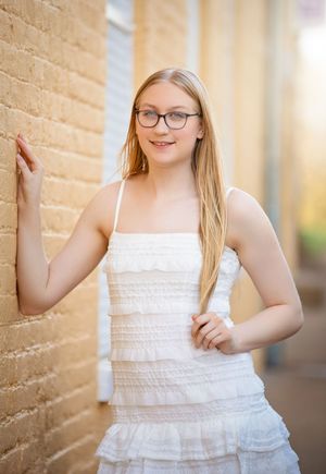 Young blonde woman in white ruffled dress and glasses leaning against brick wall outdoors.