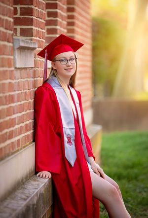 Young woman in red graduation cap and gown leaning against brick wall outdoors for senior portrait photo.