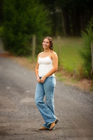 Young woman in white lace top and wide-leg jeans smiling on a rural road surrounded by green trees.