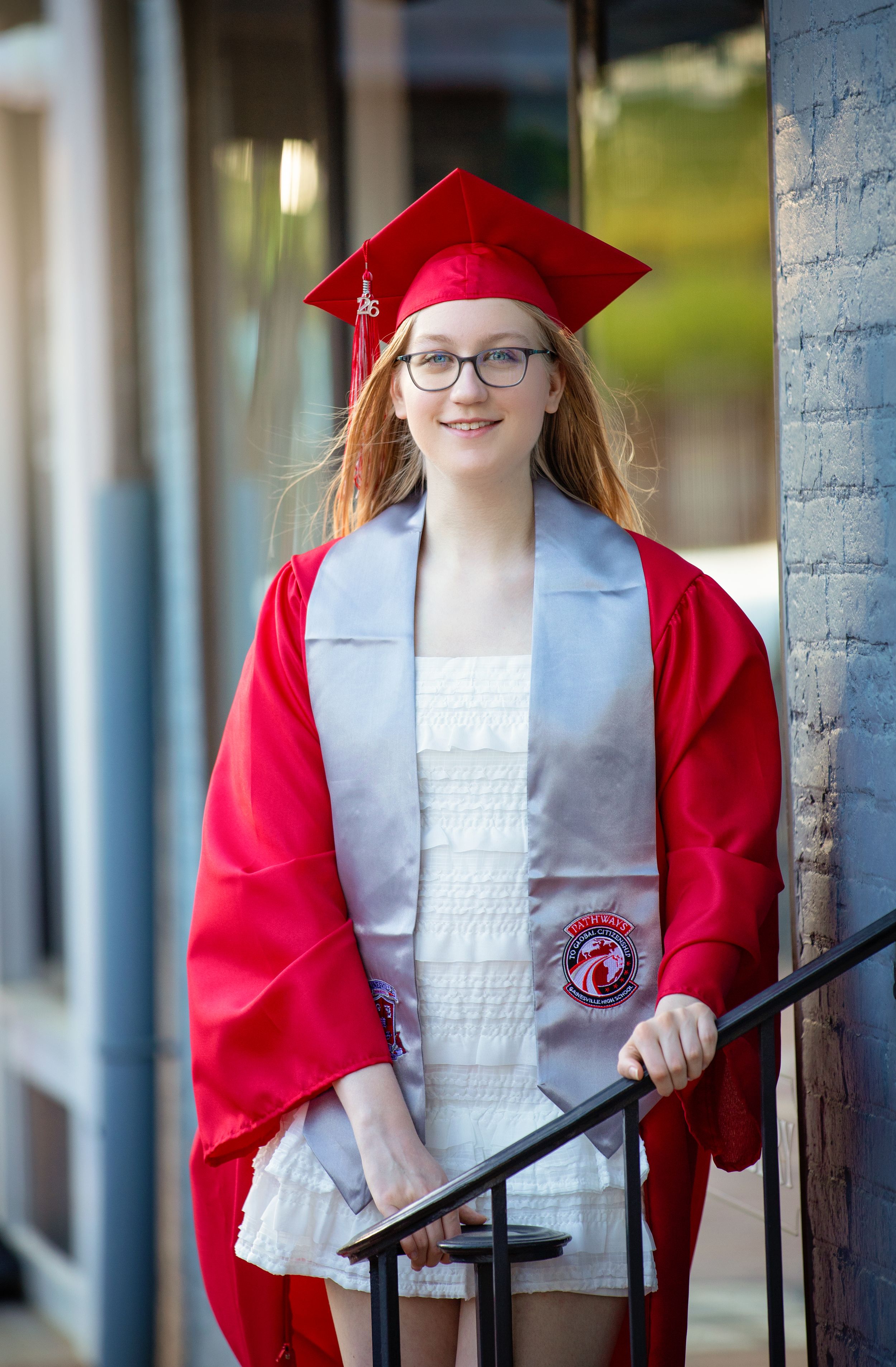 Young woman in red graduation cap and gown with silver stole smiles while holding a railing outdoors.