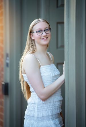 Smiling teenage girl with long blonde hair and glasses wearing a white ruffled dress, posing by a door.