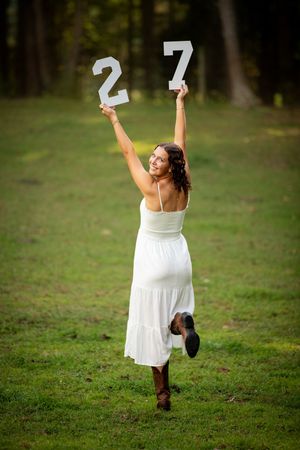 Woman in white dress and cowboy boots holds up numbers 2 and 7 outdoors in a green field.