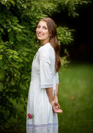 A white lace summer dress with delicate trim is showcased against lush green foliage in an outdoor setting.