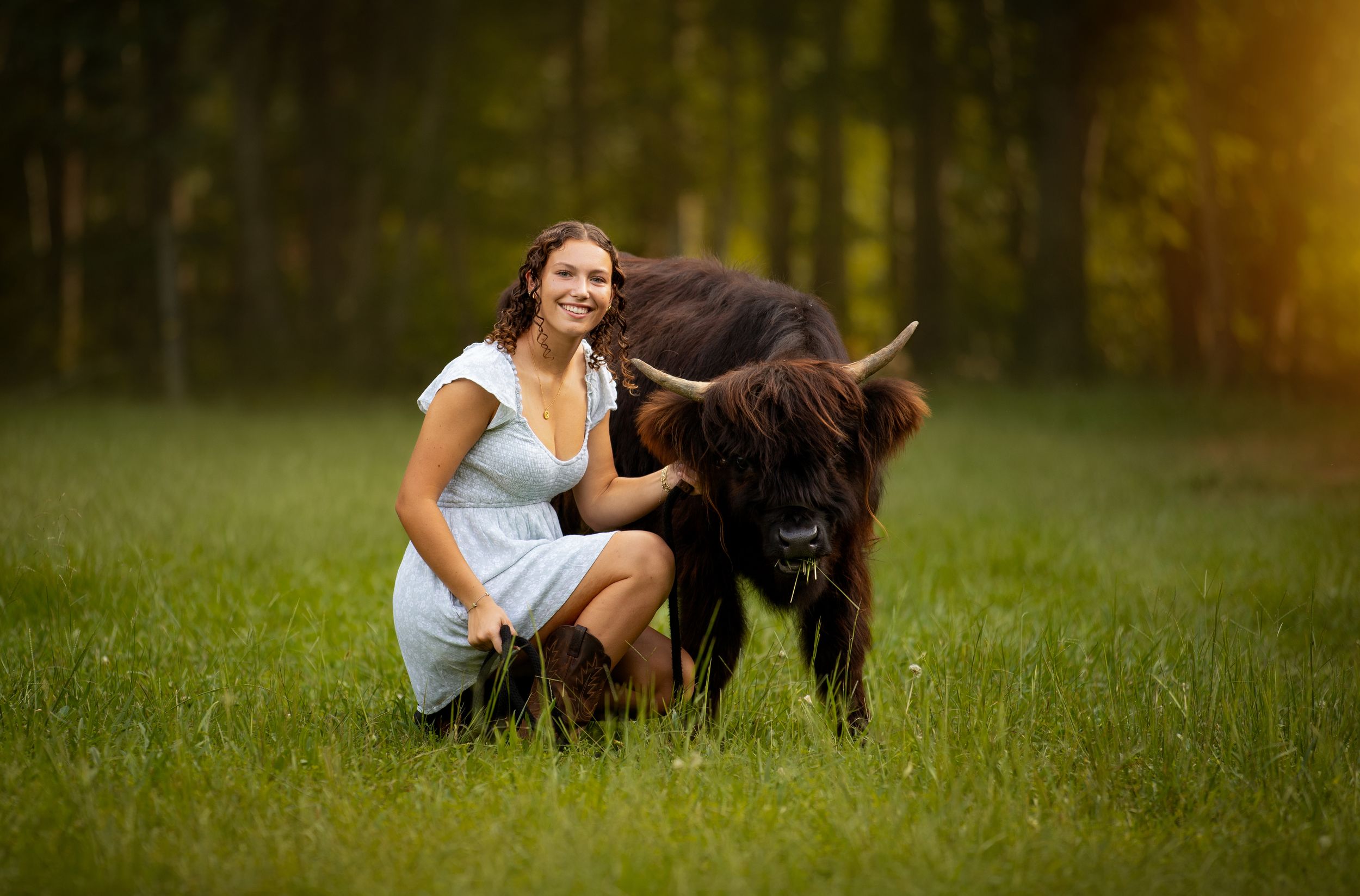 A smiling young woman in a white dress kneels beside a young highland cattle calf in a lush green meadow.