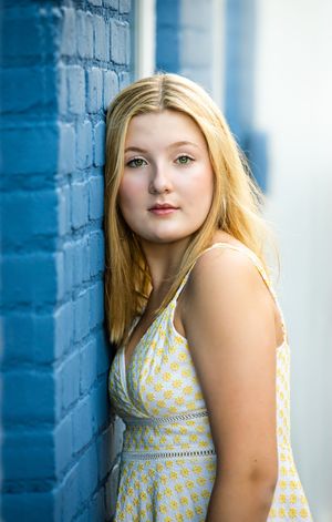 Portrait of a person in a yellow and white patterned dress against a bright blue brick wall.