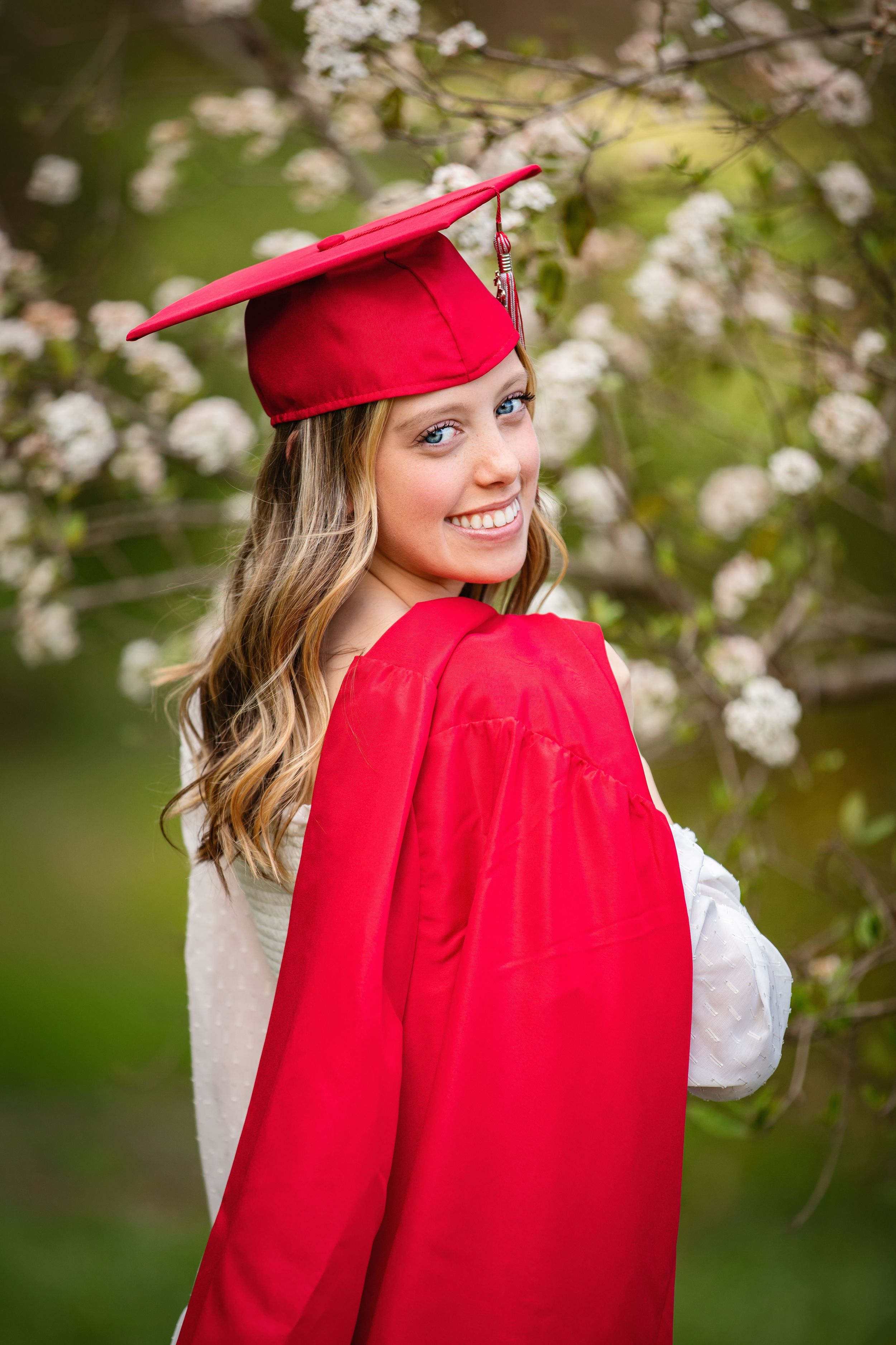 Graduation portrait in red academic regalia and cap against blooming cherry blossoms in spring.