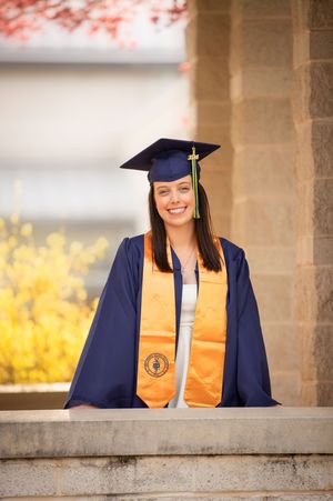 Graduate in navy cap and gown with orange stole poses in series of photos along stone wall outdoors.
