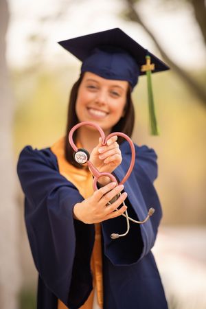 Graduate in navy blue cap and gown holds up a stethoscope in a celebratory pose outdoors.