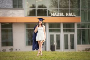 Graduate in blue cap and gown walks across campus in front of Hazel Hall building during commencement ceremony.